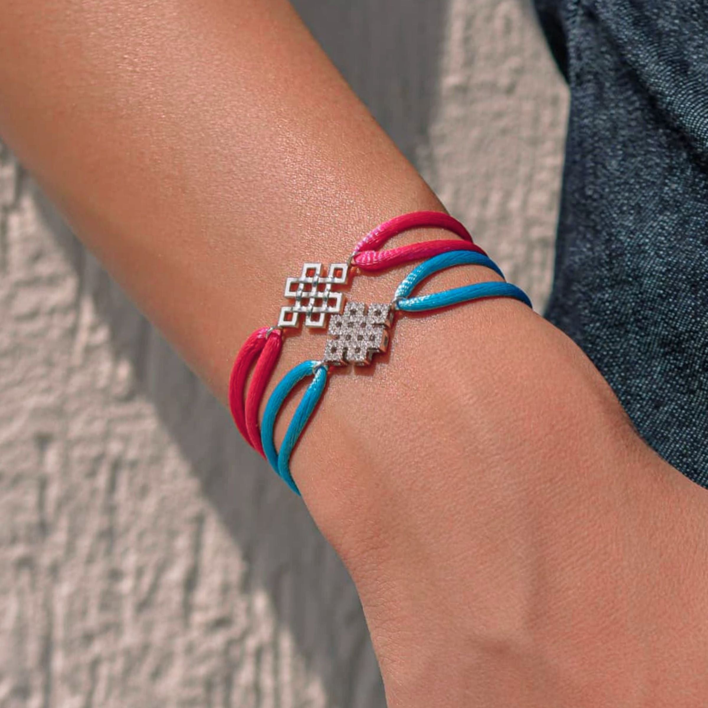 Bracelet with red and blue cords and a silver clasp on a person's wrist against a textured wall.