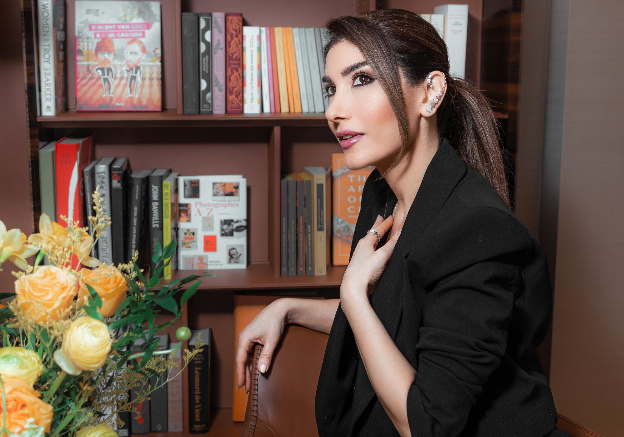 Josette Leon in a black outfit sitting in front of a bookshelf wearing her jewelry
